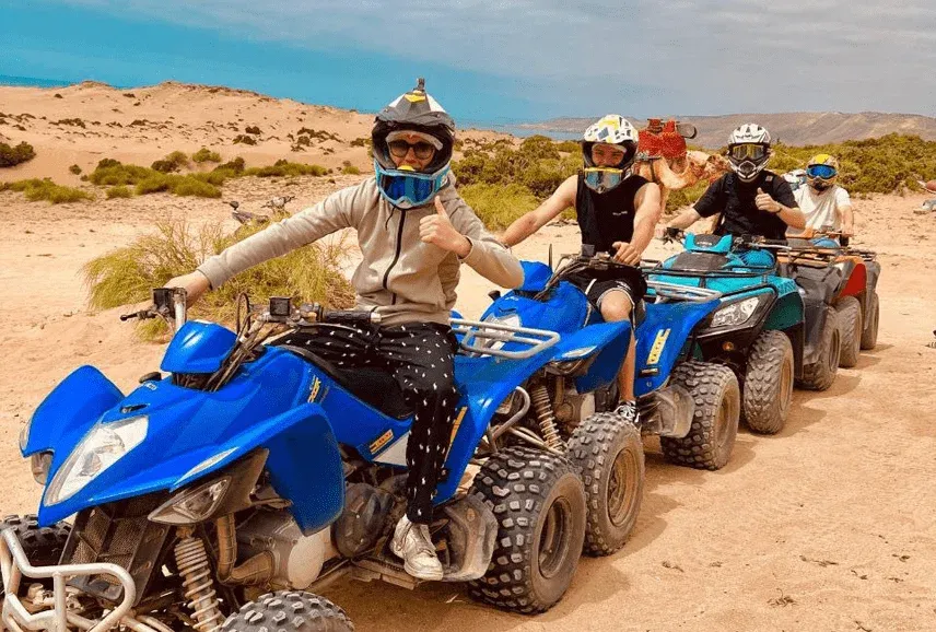 A line of riders in helmets on blue quad bikes in the scrub and sand behind the coast