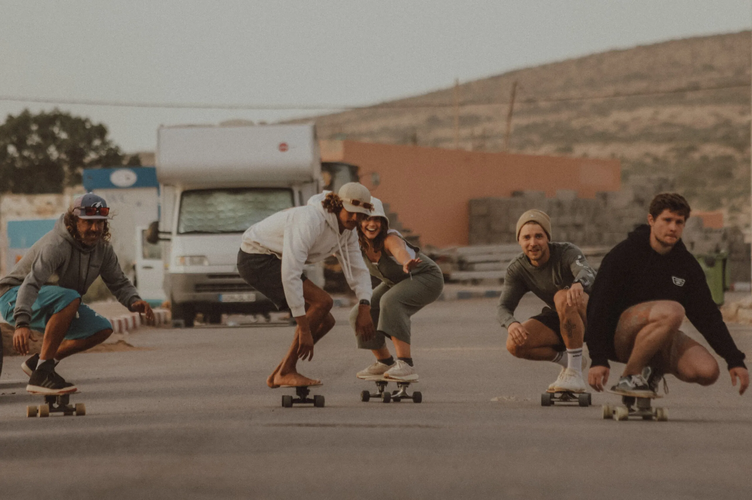 Five riders crouched low on surf-skates rolling down a road at golden hour, a camper van and the hills behind them