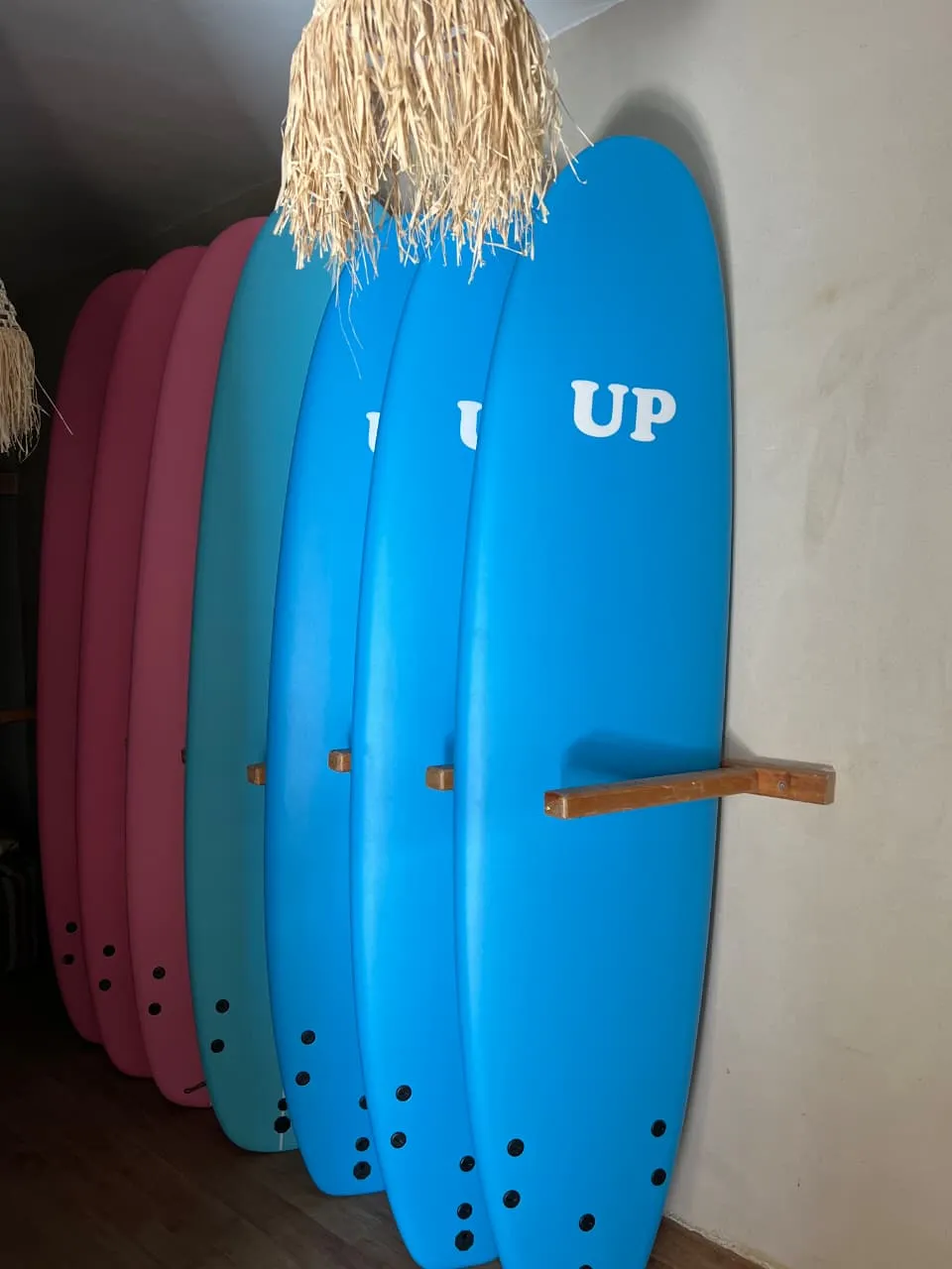 Pink and blue soft-top surfboards racked against the wall under a raffia shade at the surf house