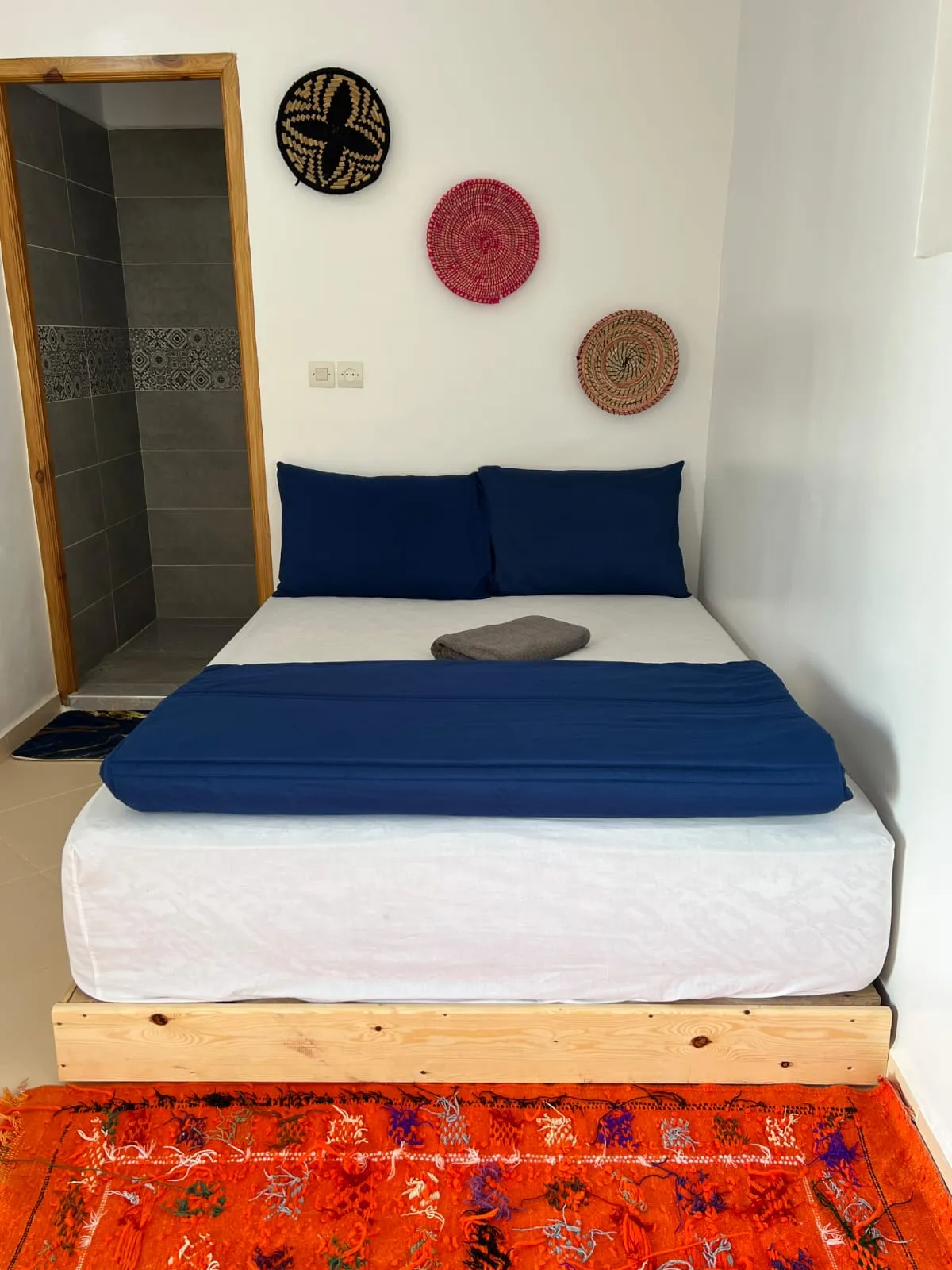 A private room at the surf house: navy bed linen, woven Berber wall discs, an orange kilim rug and an ensuite shower