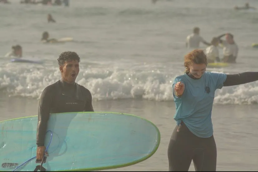A coach carrying a longboard out of the shallows beside a surfer in a blue rash vest, a busy line-up behind them