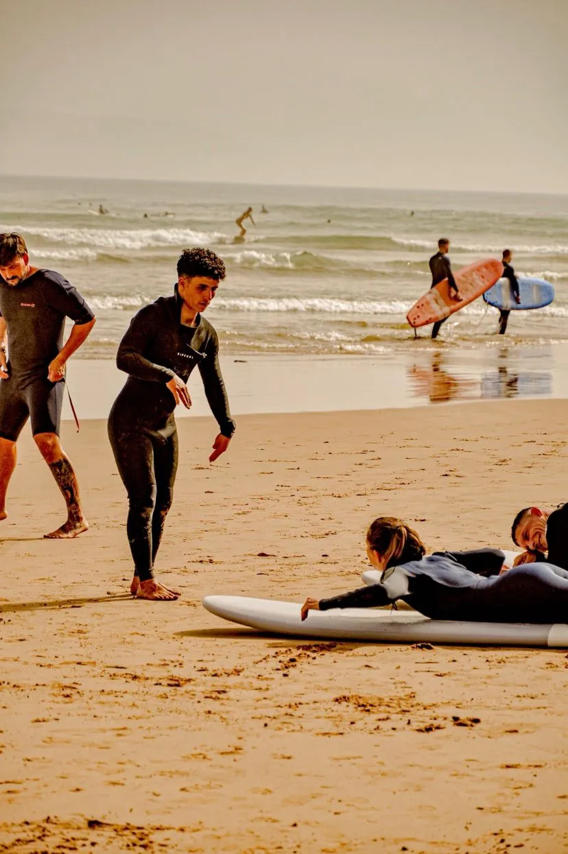 A coach talking a surfer through the pop-up on a board on the sand, others carrying boards down to the water behind