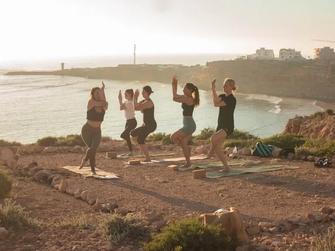 Five people holding chair pose on mats on a clifftop at golden hour, a Moroccan coastal village and the bay behind them