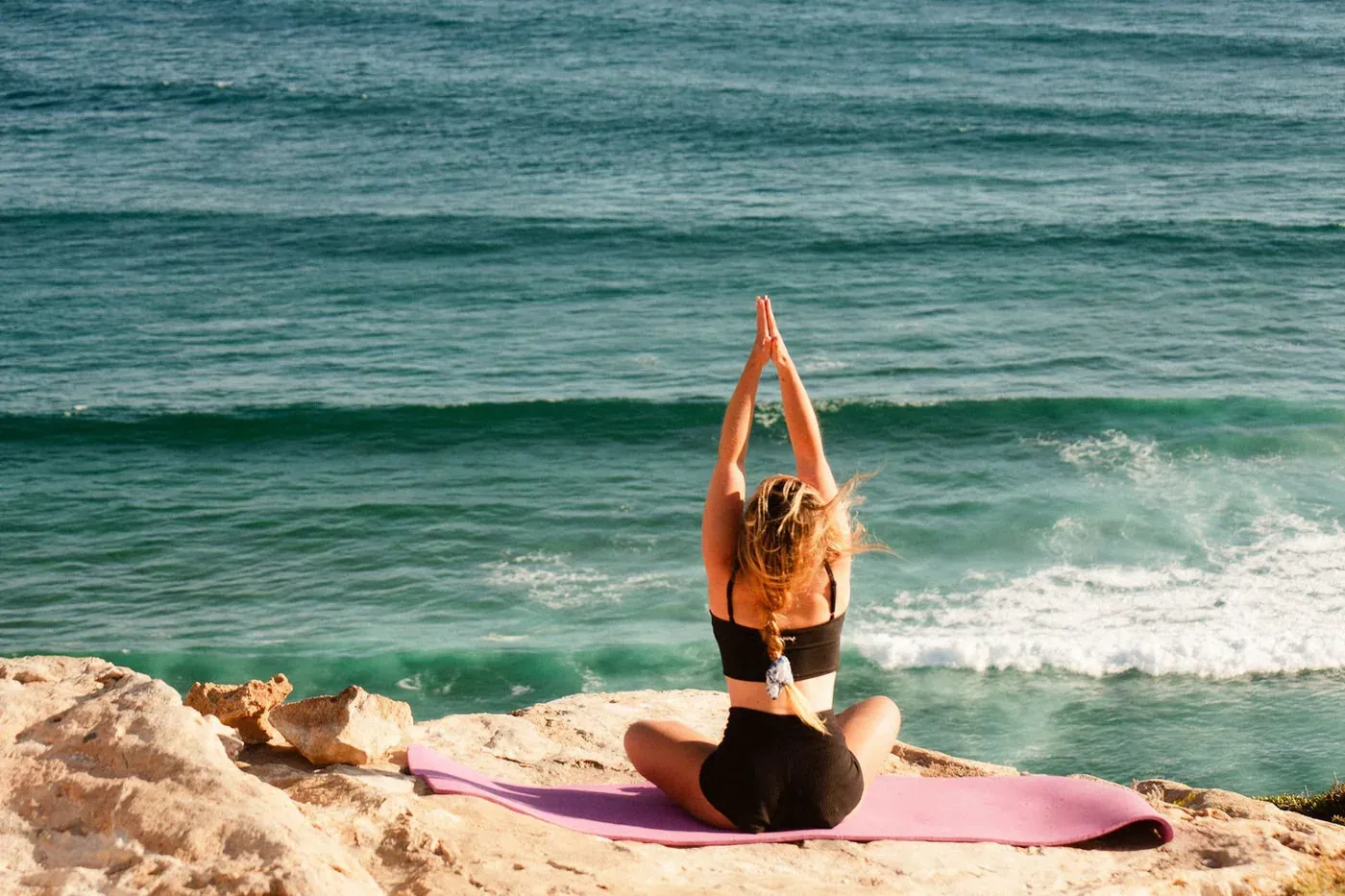 A woman sitting in yoga posture on a mat on a rock above a turquoise ocean